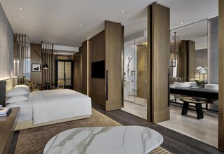 A modern hotel room with a large bed, a sitting area with a table and chairs, a bathroom with a large mirror, and a view of the city.