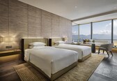 A modern hotel room with two beds, a sofa, a chair, and a large window offering a city view.