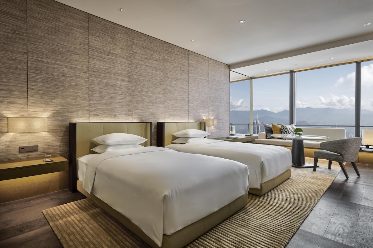 Una habitación de hotel moderna con dos camas, un sofá, una silla y un amplio ventanal con vistas a la ciudad.