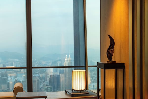 A room with a large window, a table with a lamp, and a decorative piece on the wall.