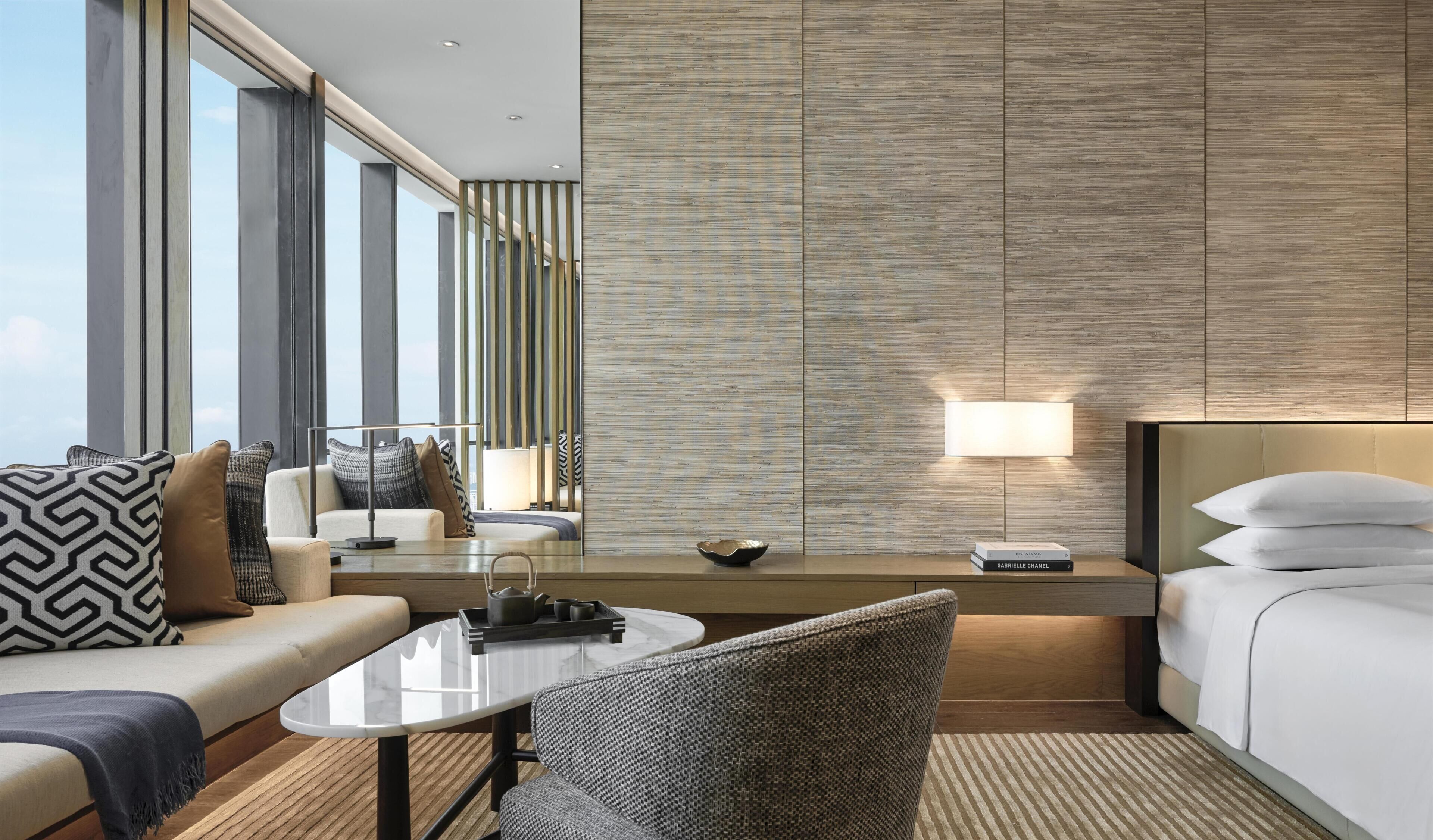 A modern hotel room with a sofa, a coffee table, a chair, a bedside table with a lamp, and a large window.