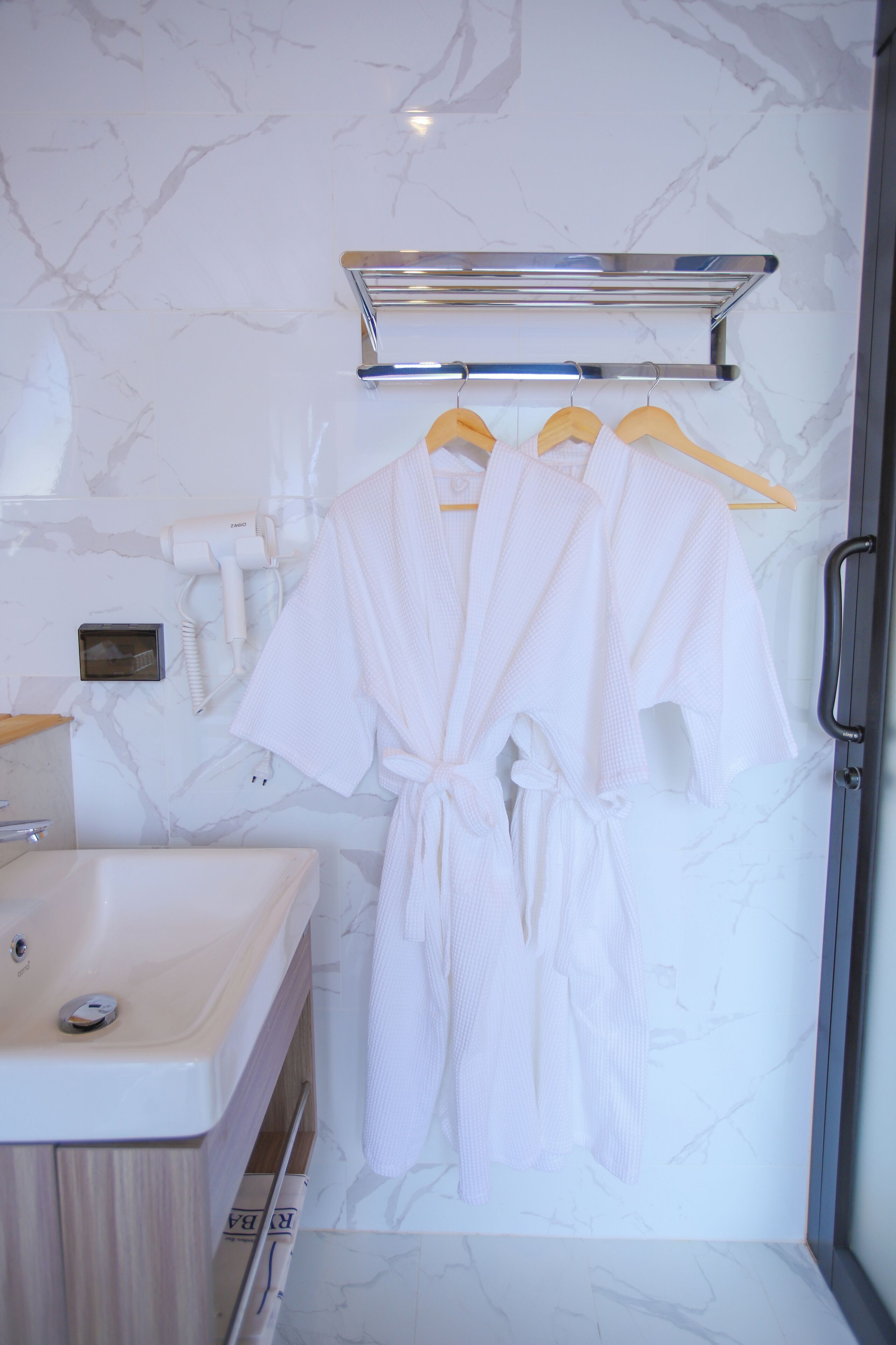 A bathroom with a marble wall, a sink, a hairdryer, and two white robes hanging on a rack.