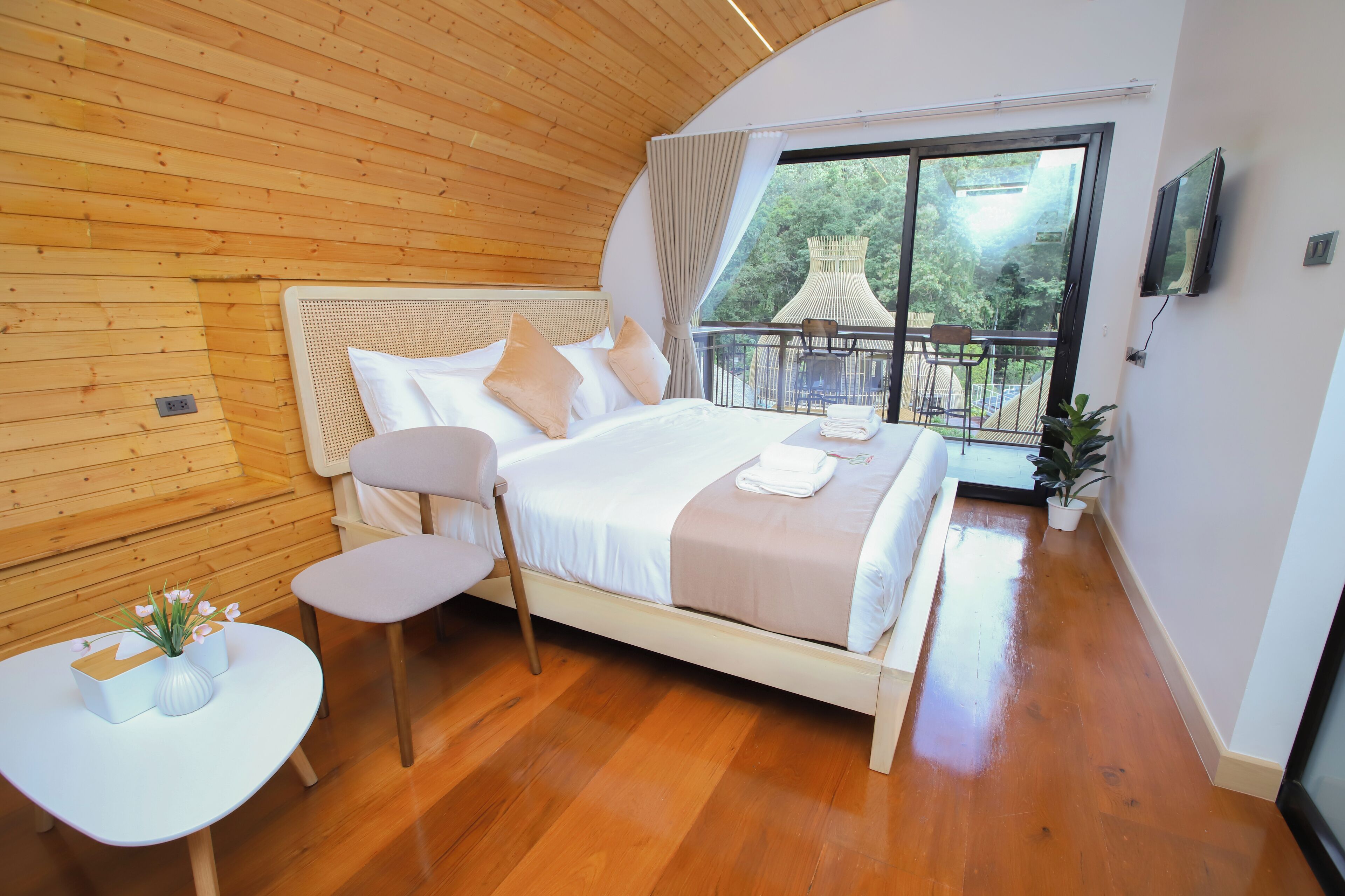 A bedroom with a wooden ceiling, a large bed with white linens, a small table with a vase, and a balcony with a chair and table.