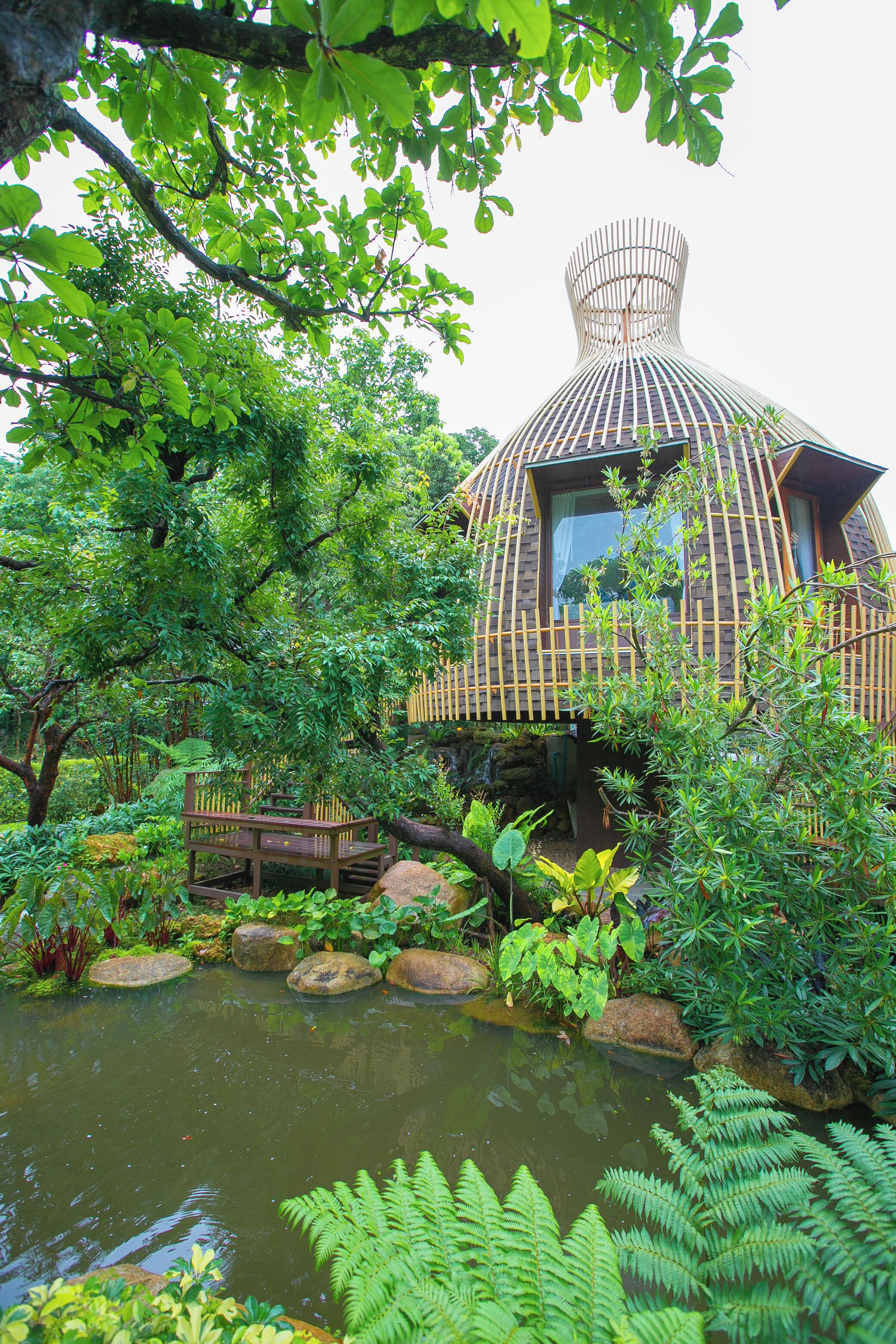 A house with a unique conical roof, surrounded by lush greenery and a pond.