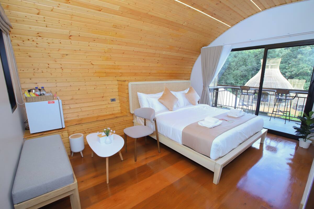A bedroom with a wooden ceiling, a bed with white bedding, a small table with chairs, a bench, and a view of a traditional building through large windows.