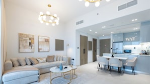 A modern living room with a sofa, coffee table, and dining area with chairs. There is a kitchen area with appliances visible.