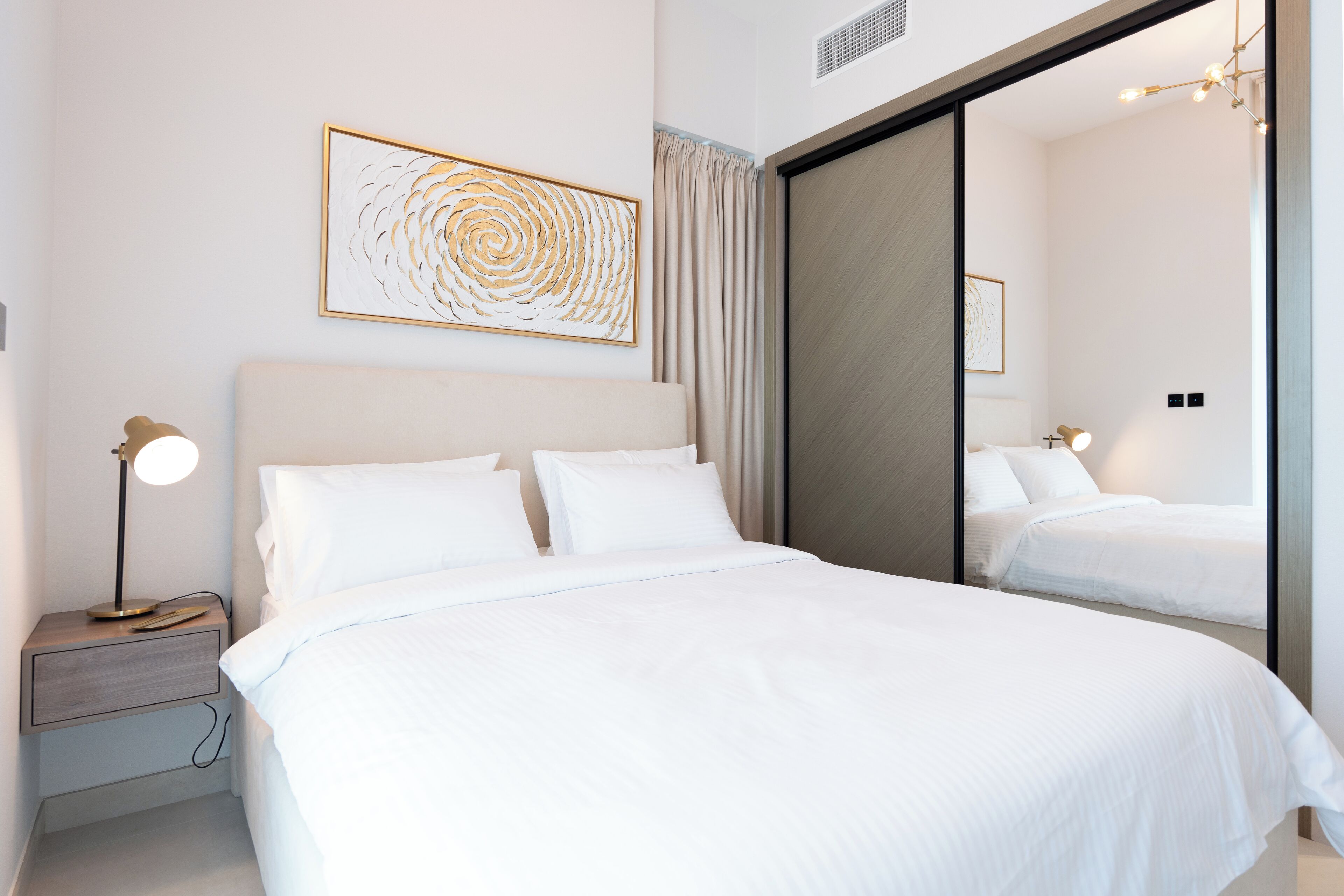 A modern hotel room with a large bed, a bedside table with a lamp, a mirror, and a wall-mounted artwork.