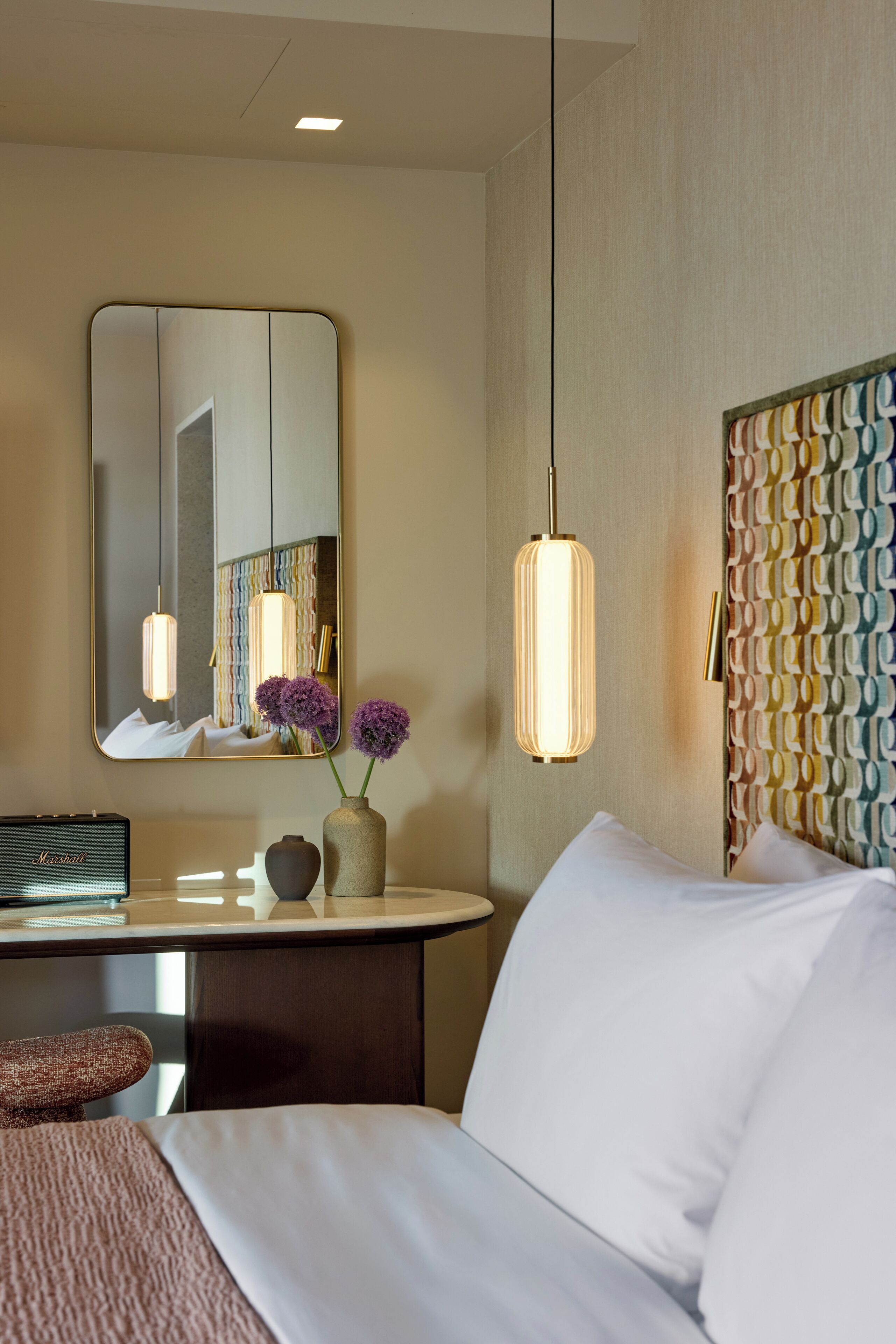 Habitación de hotel con una cama, una mesita de noche con un radio, un florero con flores, un espejo y un cabecero estampado.