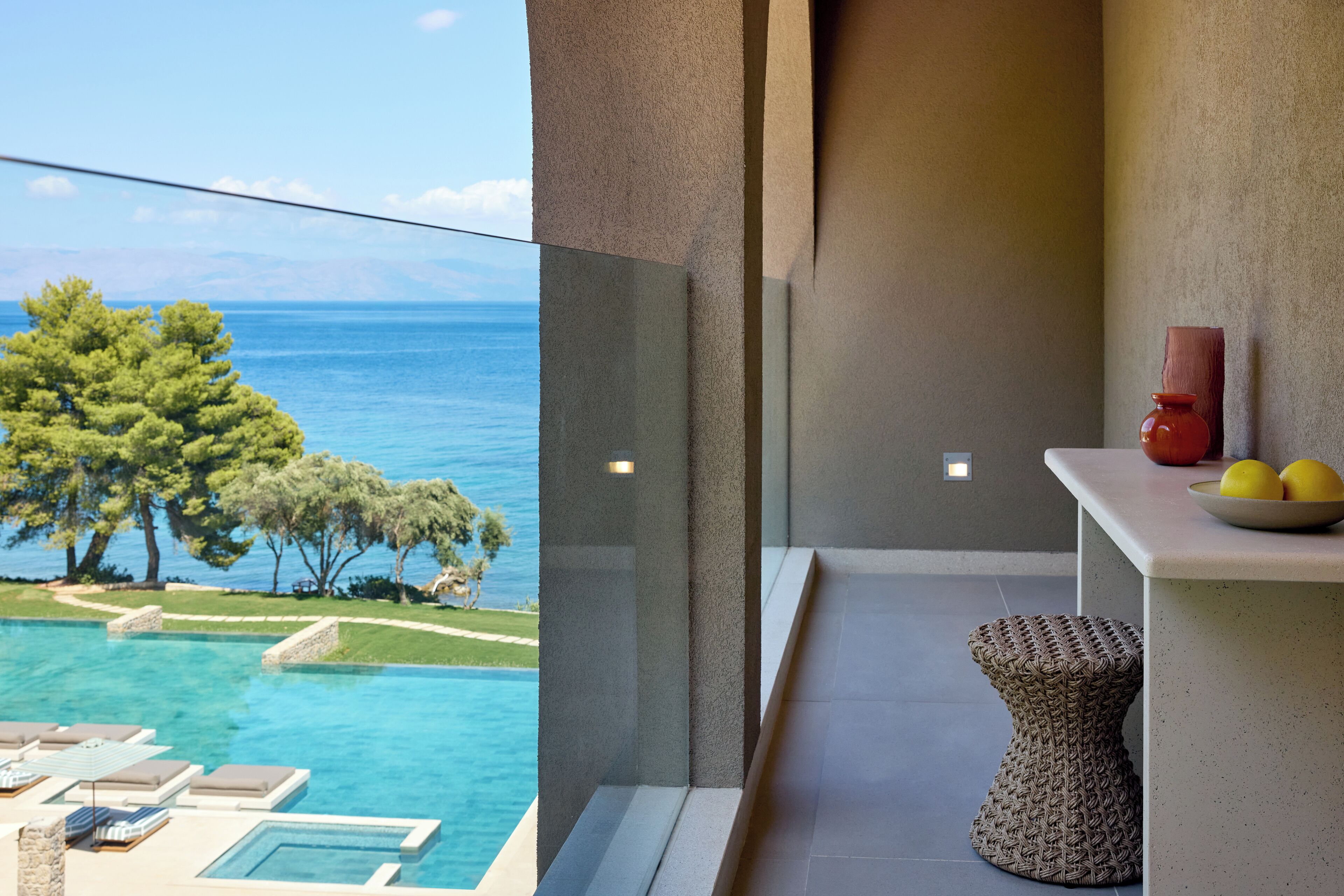 Un balcon avec vue sur la mer, une petite table garnie de fruits et un tabouret en osier.