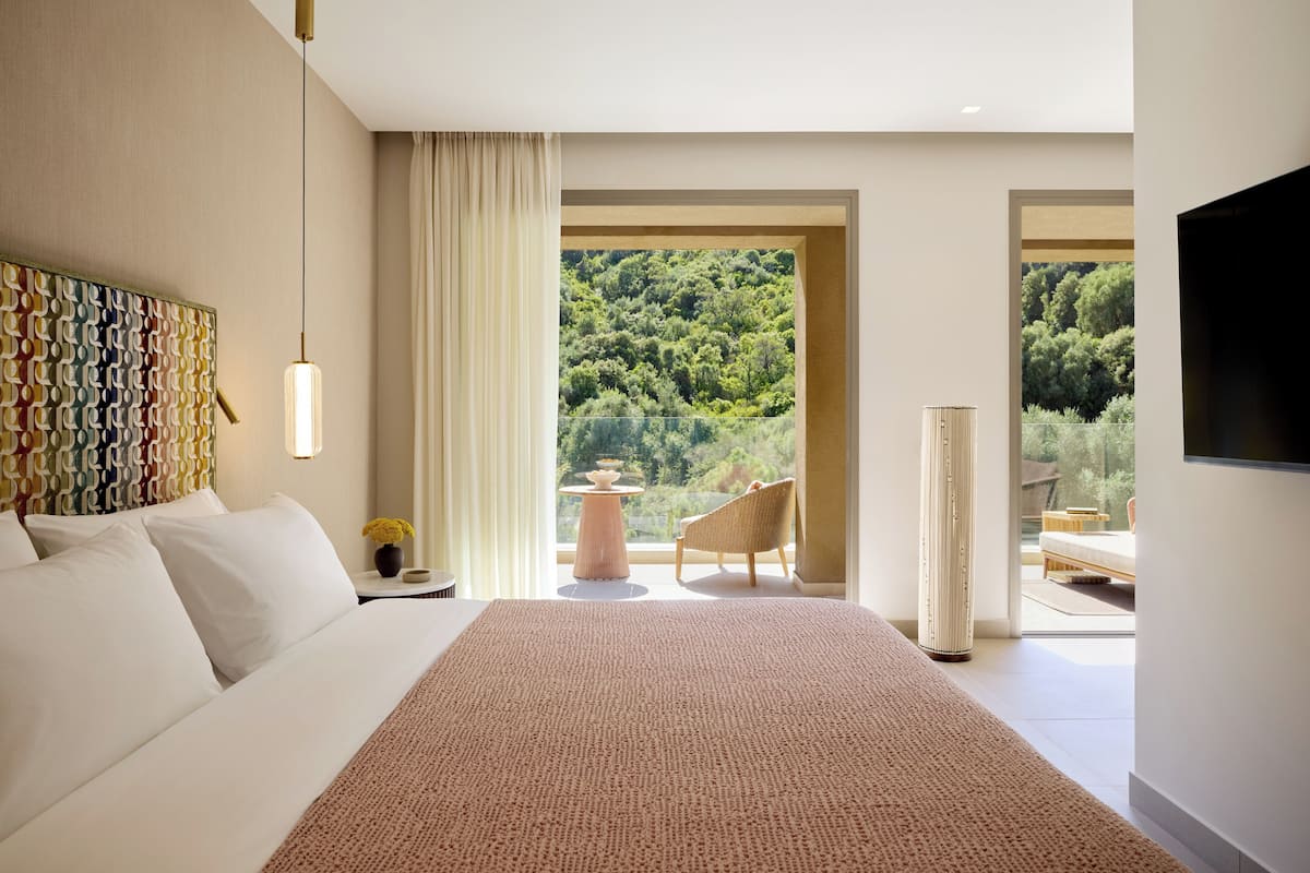 Une chambre d’hôtel moderne dotée d’un grand lit, offrant une vue sur une zone boisée, comprenant un coin salon avec une table et des chaises, et d’un téléviseur à écran plat fixé au mur.