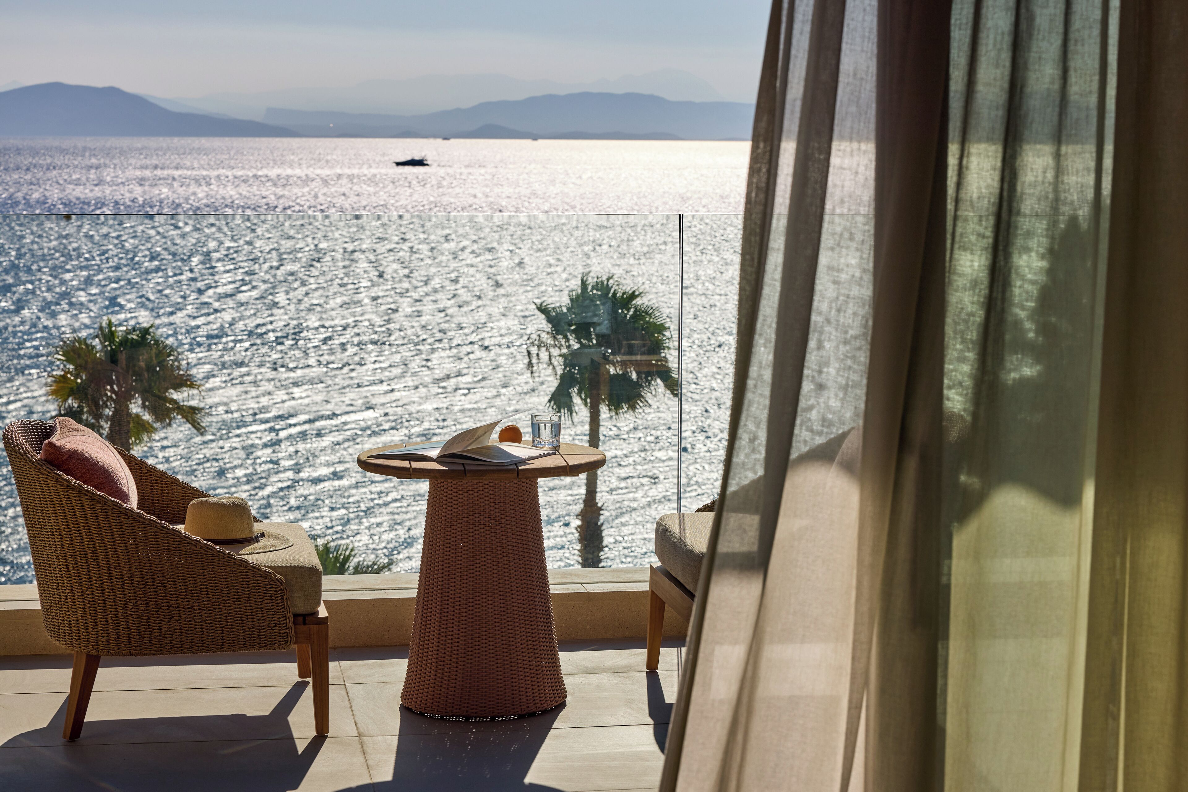 Un balcon agrémenté de meubles en osier, d’une petite table et offrant une vue sur la mer et les montagnes lointaines.