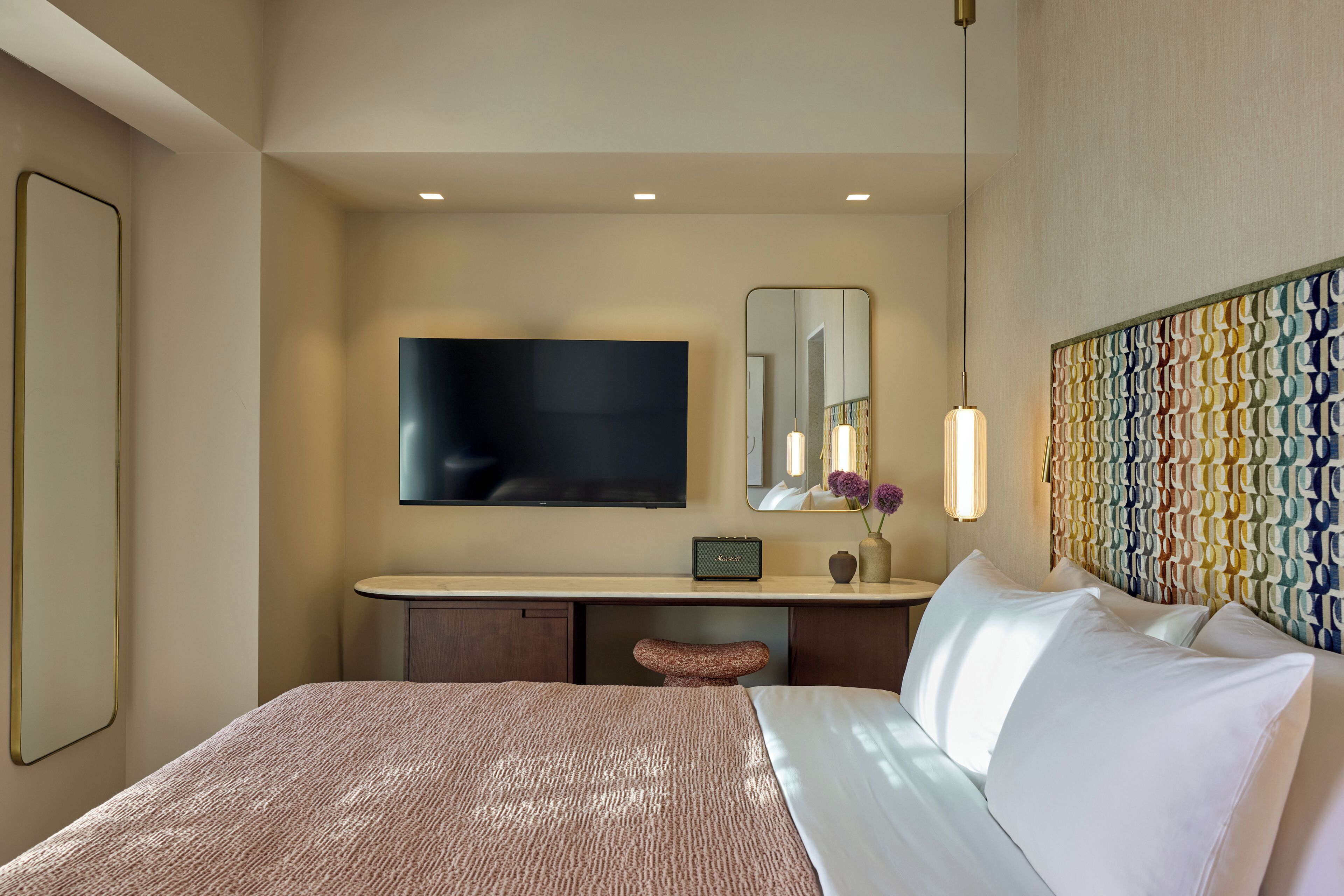 Una habitación de hotel con una cama grande, un televisor de pantalla plana en la pared, un pequeño escritorio con una silla y un panel de pared colorido.
