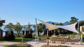 An outdoor dining area with tables and chairs, a white canopy, and a view of the sea and mountains.
