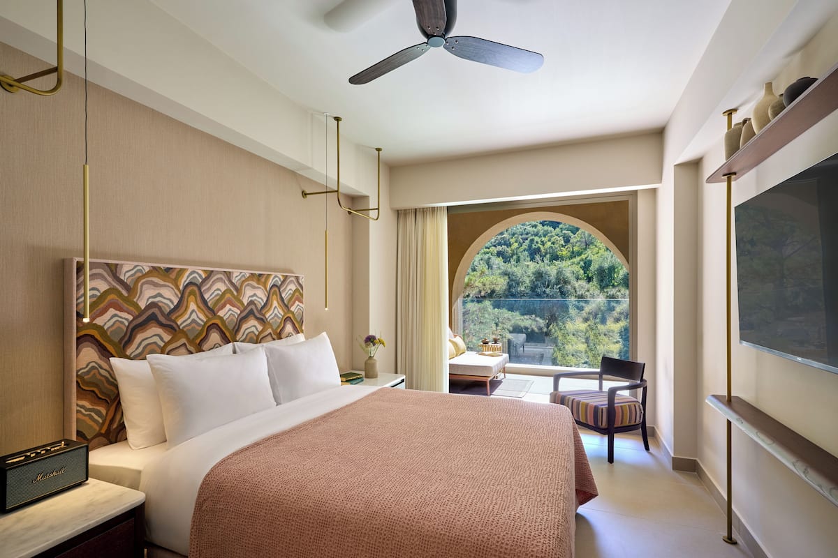 Une chambre d’hôtel équipée d’un lit, d’un bureau, d’une chaise, d’une télévision et offrant une vue sur la verdure grâce à une fenêtre en arc de cercle.