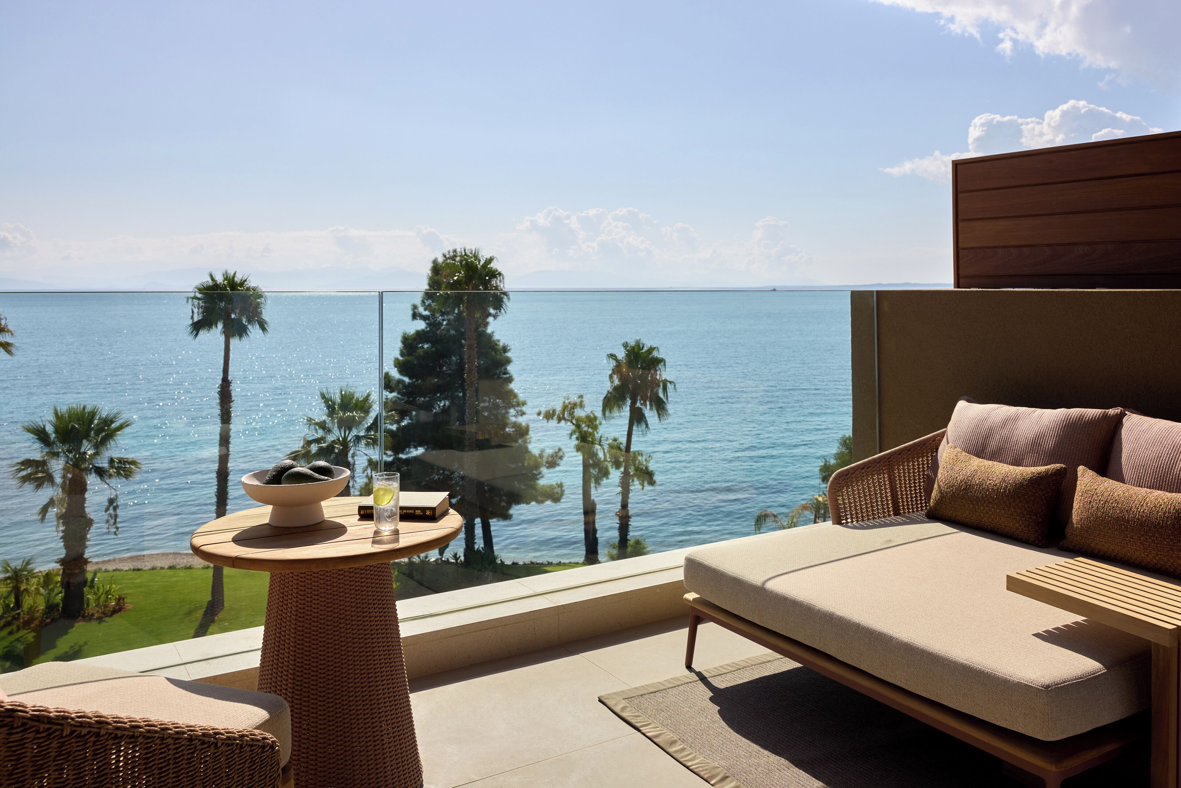 A balcony with a sofa, a small table with a bowl and a glass, and a view of the sea and palm trees.