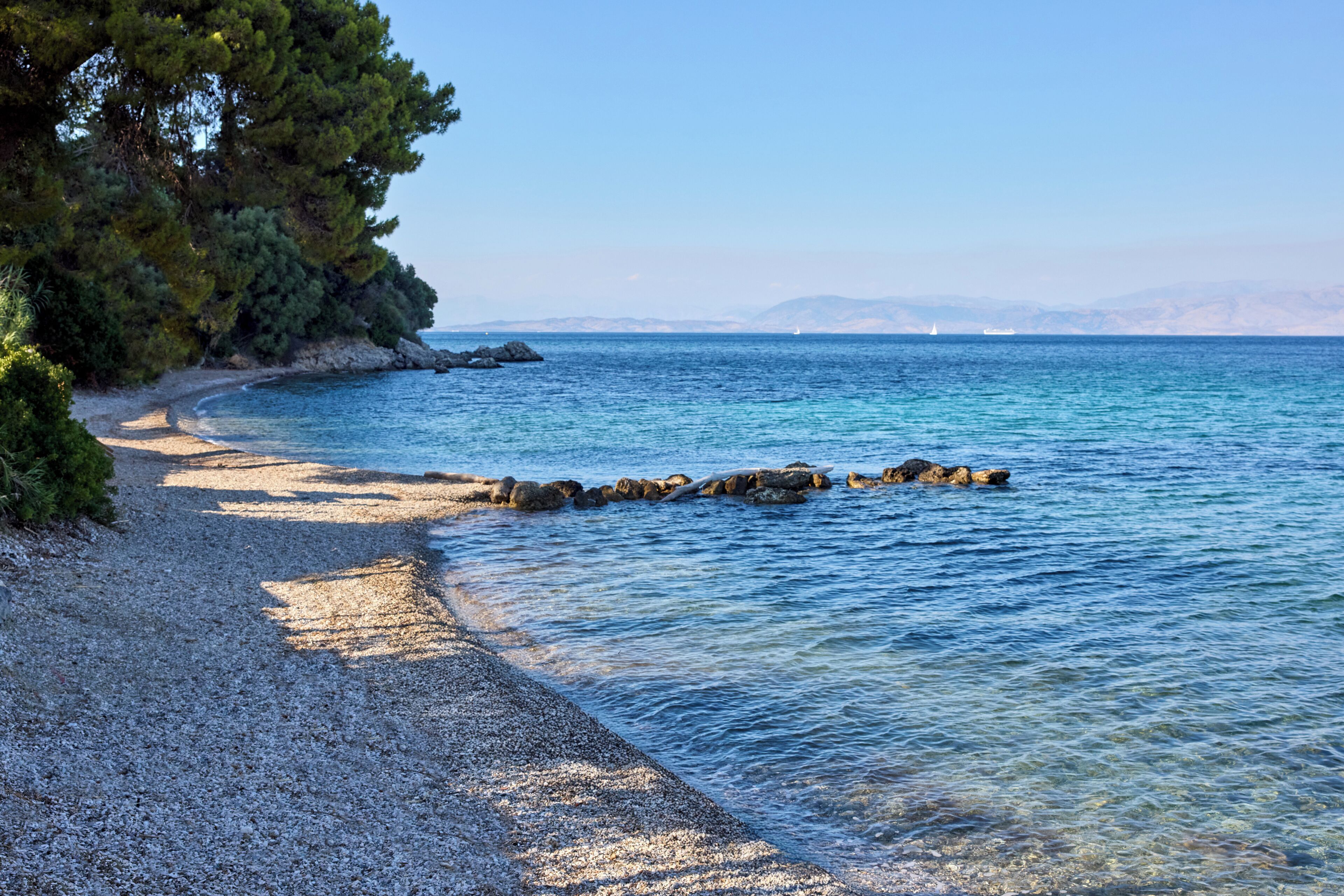 Une plage de galets bordée d’une eau d’un bleu limpide et dominée par des montagnes lointaines.