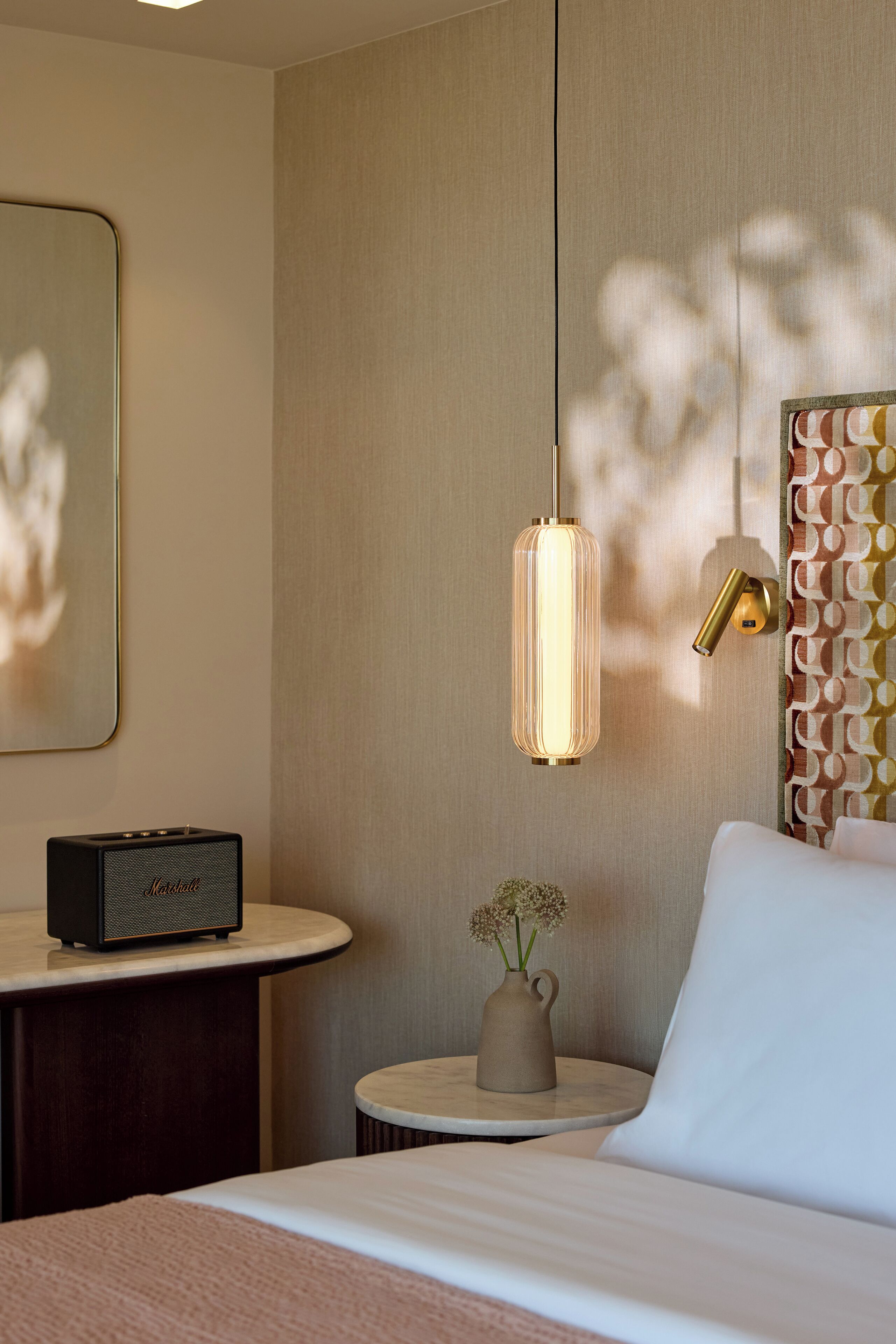 A hotel room with a bed, a bedside table with a radio, a vase with flowers, a wall-mounted lamp, and a patterned wall.