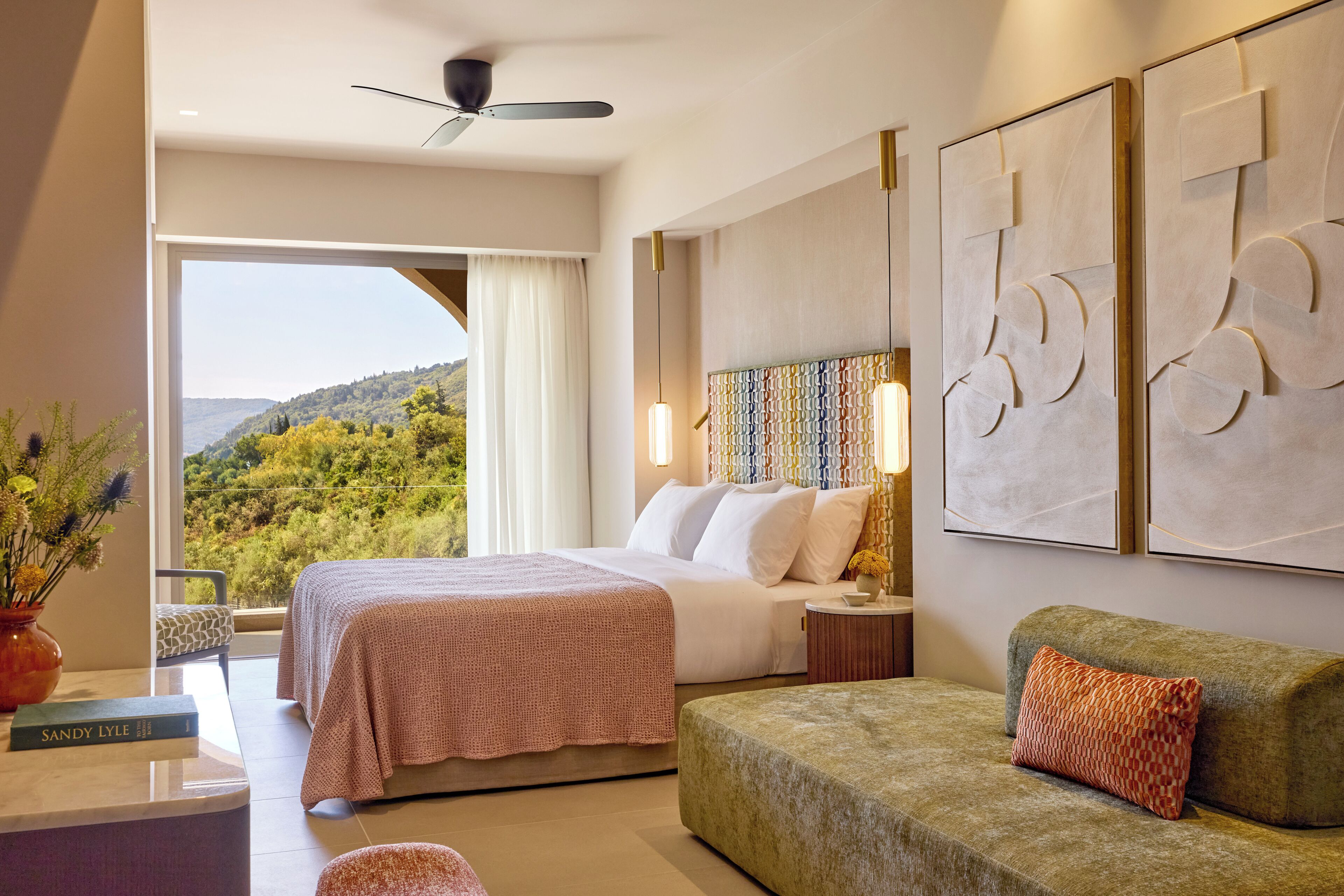 A hotel room with a bed, a sofa, a desk with a vase of flowers, and a view of the outdoors.
