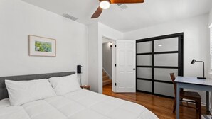 A bedroom with a bed, a desk, a chair, a ceiling fan, and a door leading to another room.