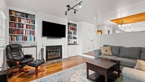 A modern living room with a fireplace, a large flat-screen TV, a leather armchair, a grey sofa, and a wooden coffee table.