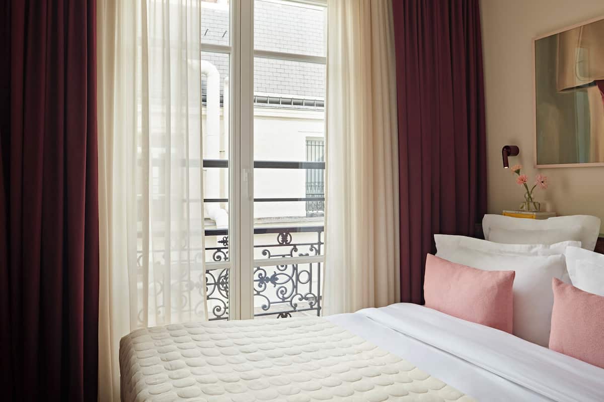 A hotel room with a bed, curtains, a window with a view of a building, and a bedside table with a lamp and flowers.