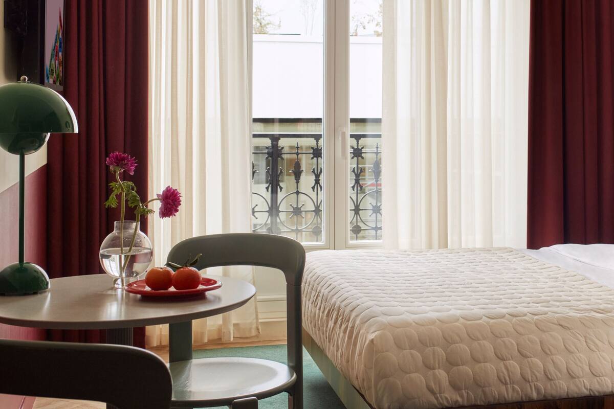 A hotel room with a bed, a round table with a vase and fruit, a chair, and a view of a balcony with a railing.