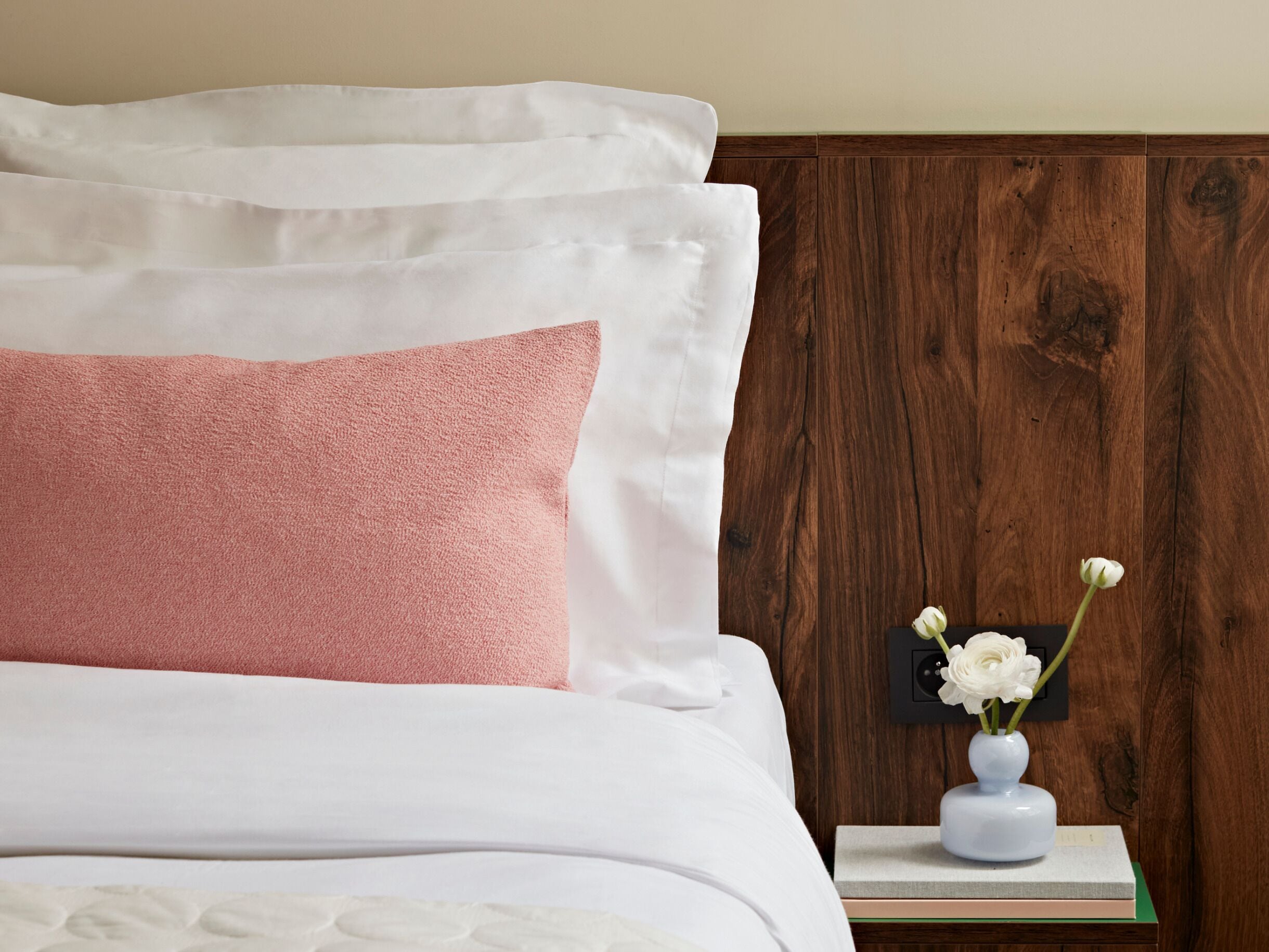 A neatly made bed with white and pink pillows, a small white vase with flowers on a bedside table, and a wooden headboard.
