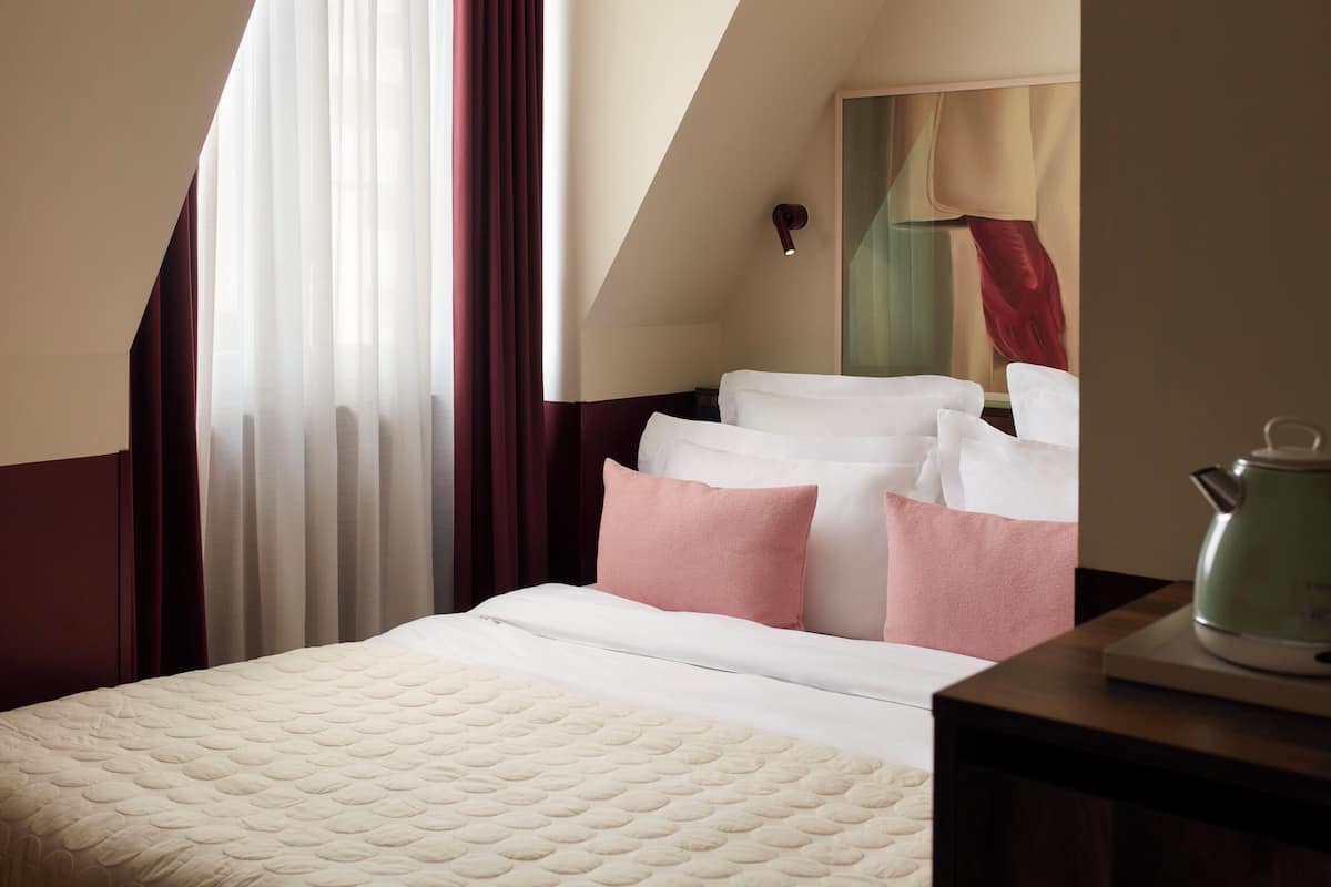 A hotel room with a bed, pillows, a nightstand, a kettle, and a painting on the wall.