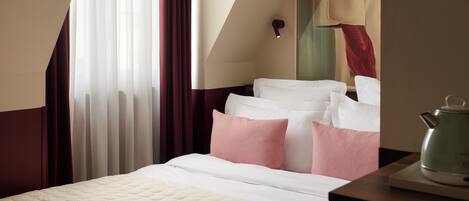 A hotel room with a bed, pillows, a nightstand, a kettle, and a painting on the wall.