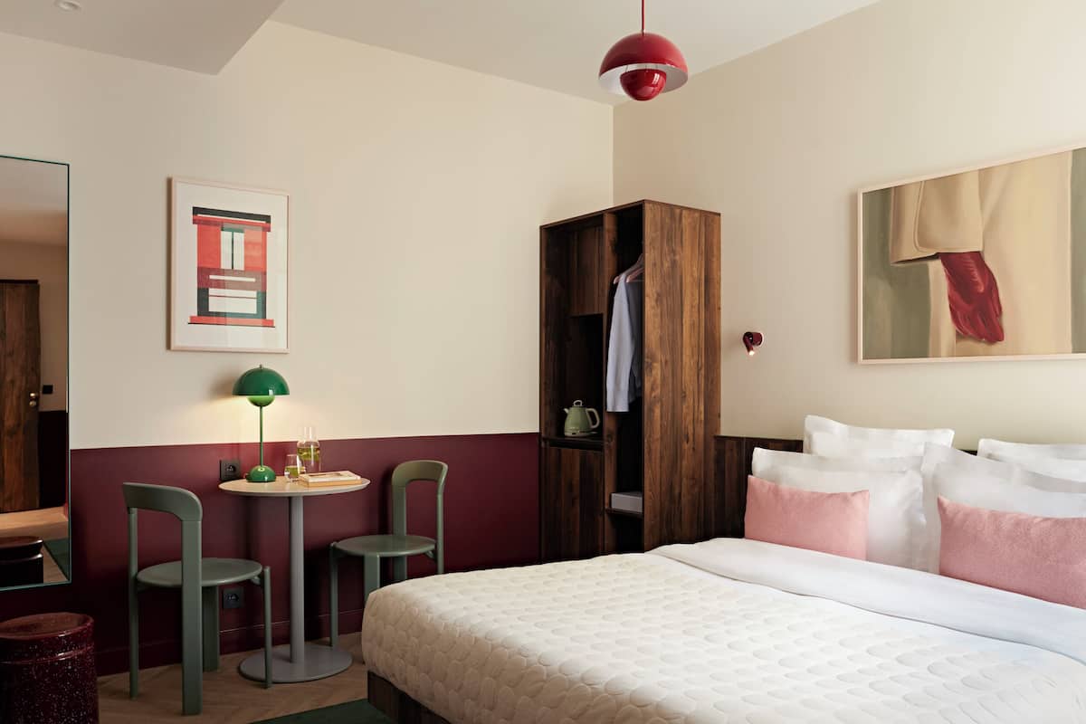 A bedroom with a large bed, a wooden wardrobe, a small table with chairs, and a painting on the wall.