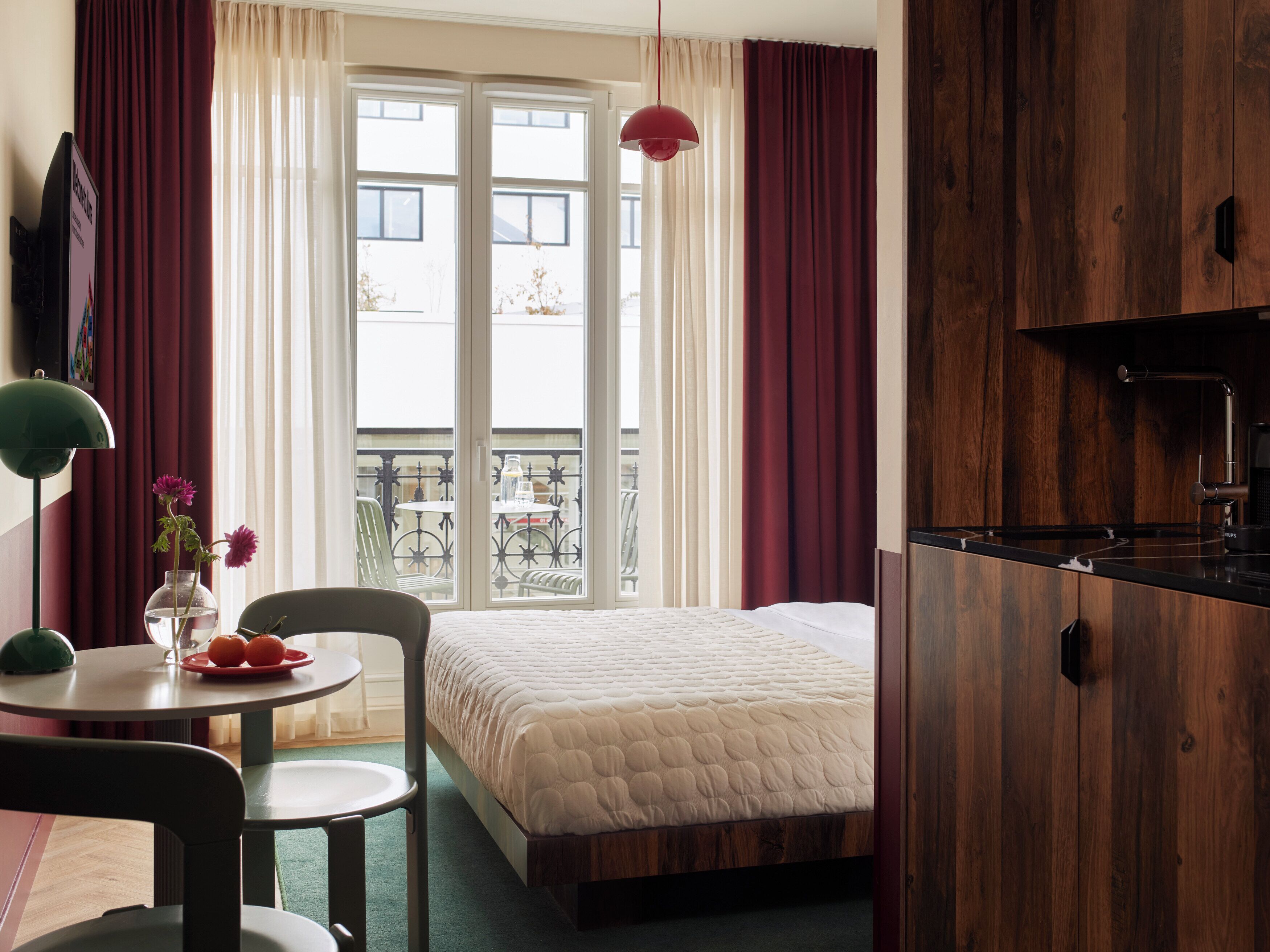 A hotel room with a bed, a dining table with chairs, a small balcony with a table and chairs, and a kitchen area.