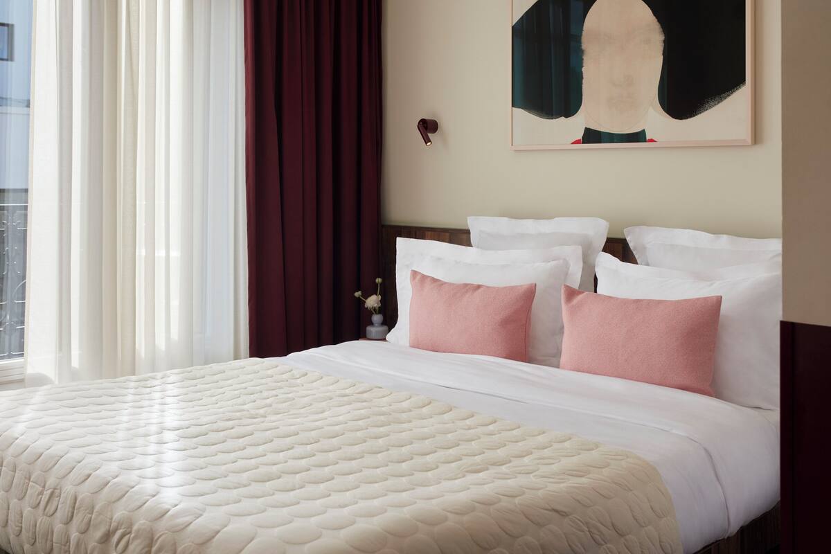 A hotel room with a large bed, white bedding, a red curtain, and a painting on the wall.