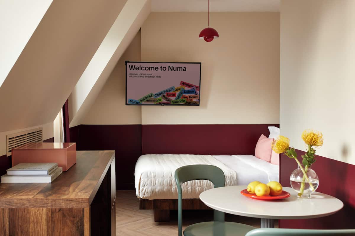 A modern hotel room with a bed, a desk, a chair, a table with fruit, and a welcome sign.