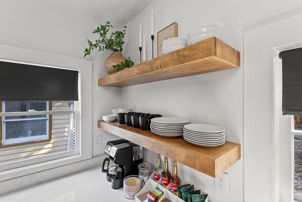 A kitchen with white walls, wooden shelves holding plates, cups, and a coffee maker. There is a window with blinds and a potted plant on the shelf.