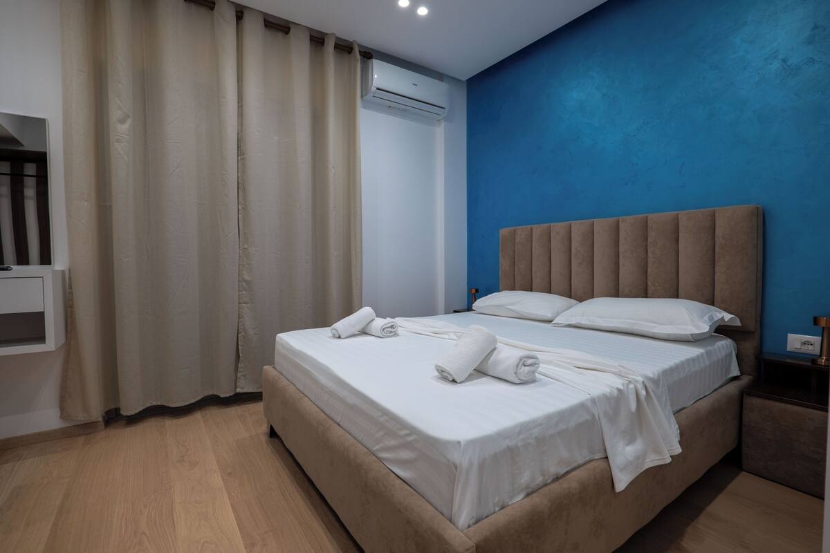 Une chambre d’hôtel moderne avec un grand lit, des rideaux beiges, un mur d’accent bleu et un appareil de climatisation.