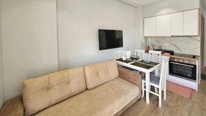 A modern living room with a beige sofa, a dining table with chairs, a flat-screen TV mounted on the wall, and a kitchen area with white cabinets and a stove. - Star&Sea Saranda (Sarandë)
