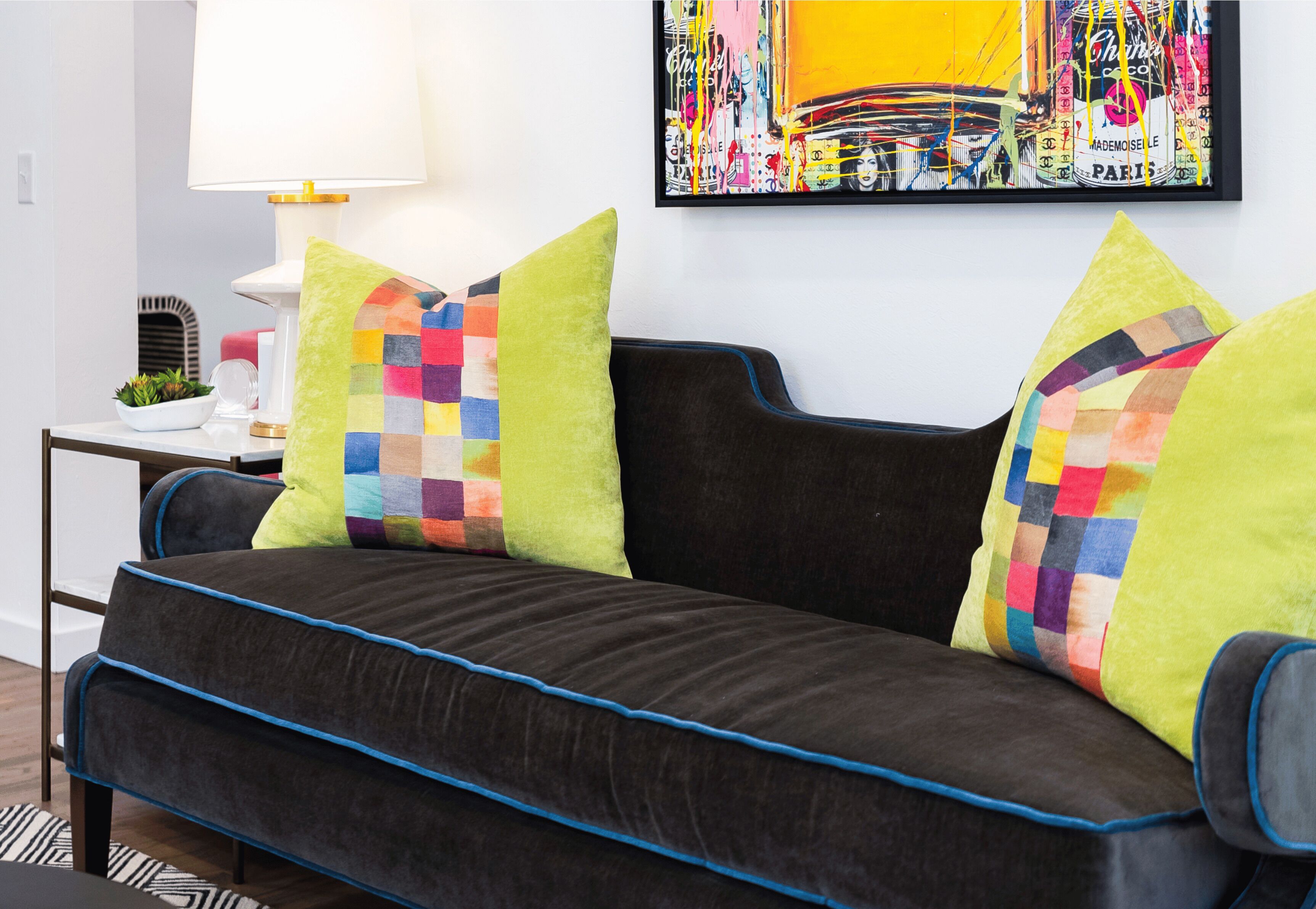 A modern living room with a dark sofa, colorful cushions, a white side table with a lamp, and a framed abstract painting on the wall.