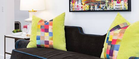 A modern living room with a dark sofa, colorful cushions, a white side table with a lamp, and a framed abstract painting on the wall.