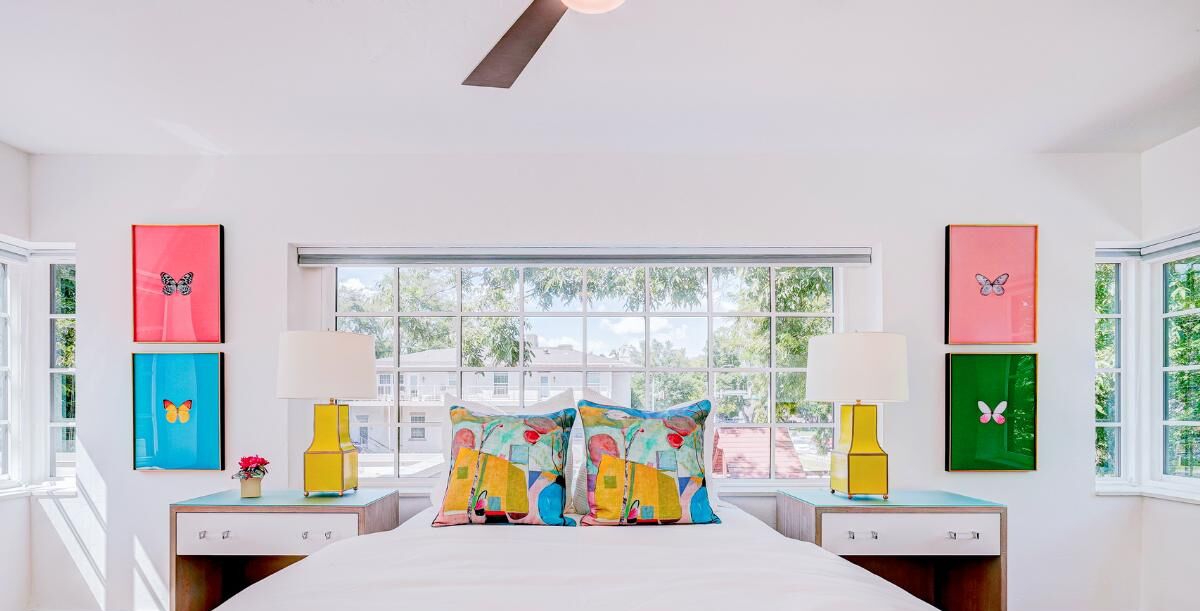 Un dormitorio con una cama grande, dos mesitas de noche, un ventanal con vistas a edificios y coloridas obras de arte en la pared.