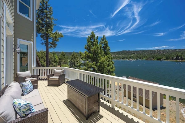 Una terraza con mobiliario exterior, una mesita y una barandilla con vistas a un lago rodeado de árboles y colinas.