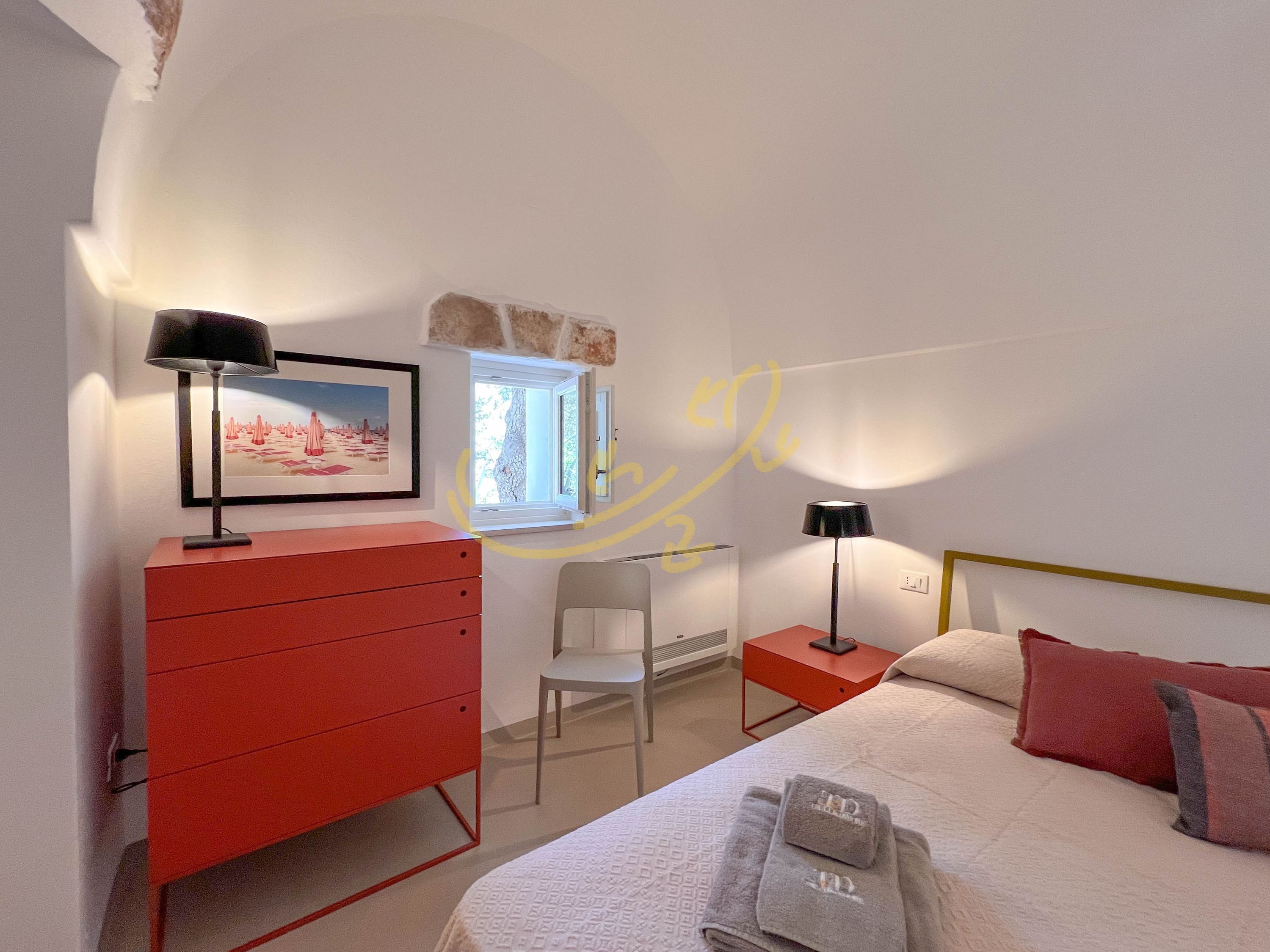 A bedroom with a bed, a red dresser, a desk, a chair, a window, and a picture on the wall.