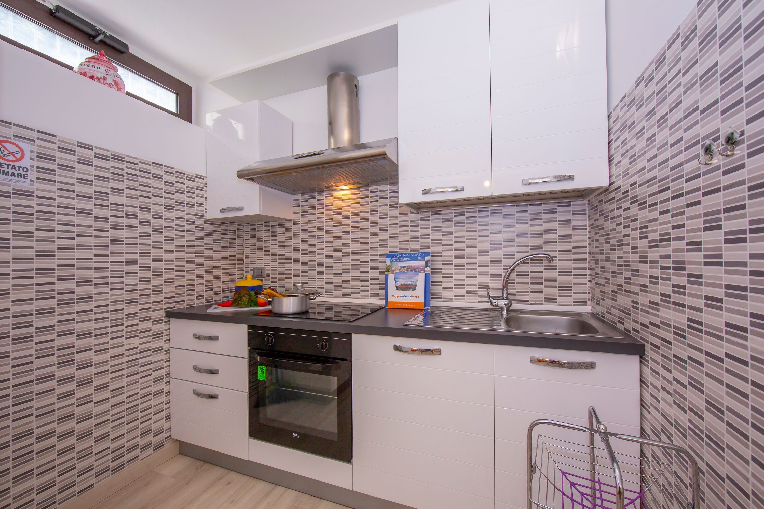 A compact kitchen with white cabinets, a stainless steel range hood, a built-in oven, and a tiled backsplash.