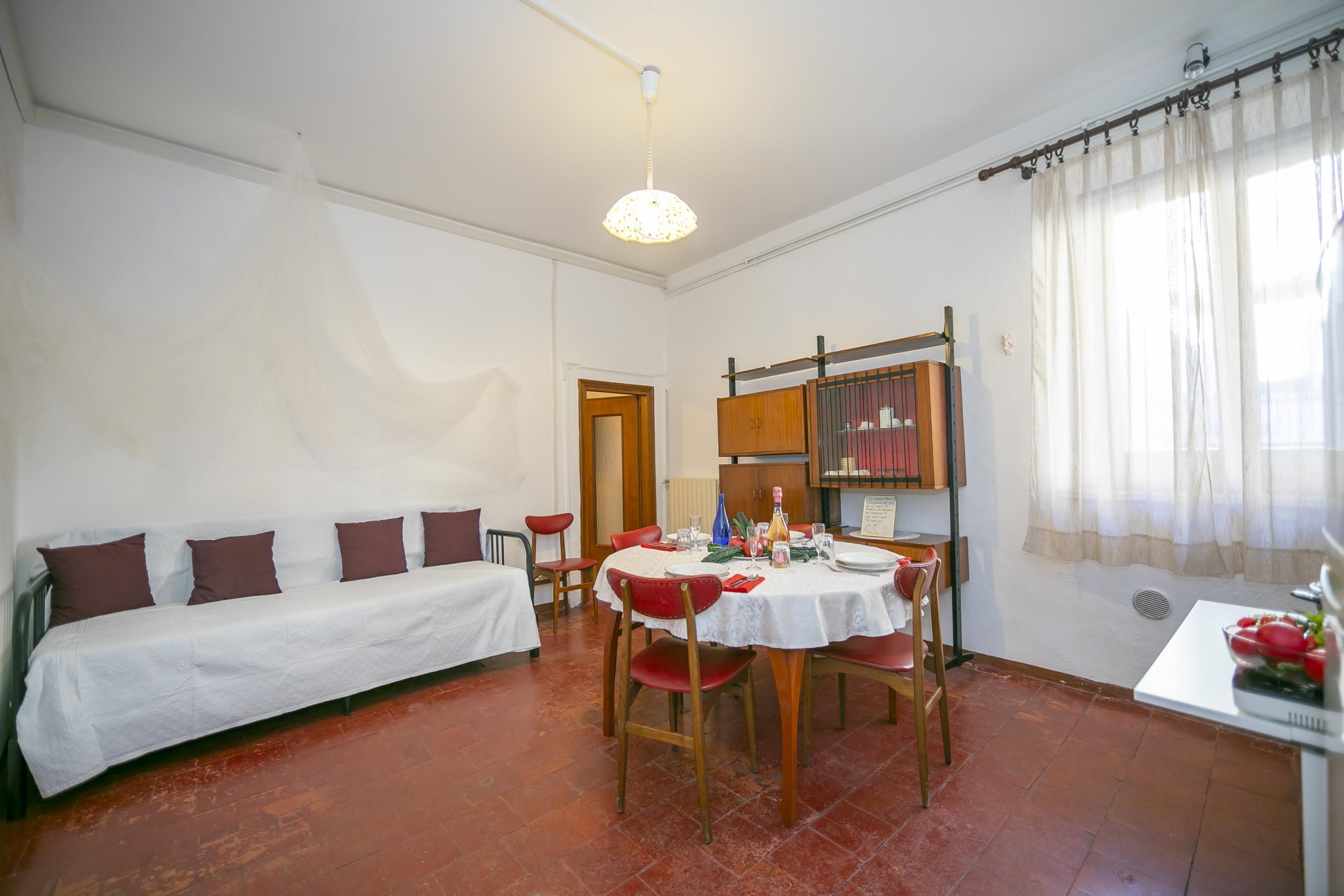 A room with a bed, a dining table with chairs, a ceiling light, a window with curtains, and a radiator.