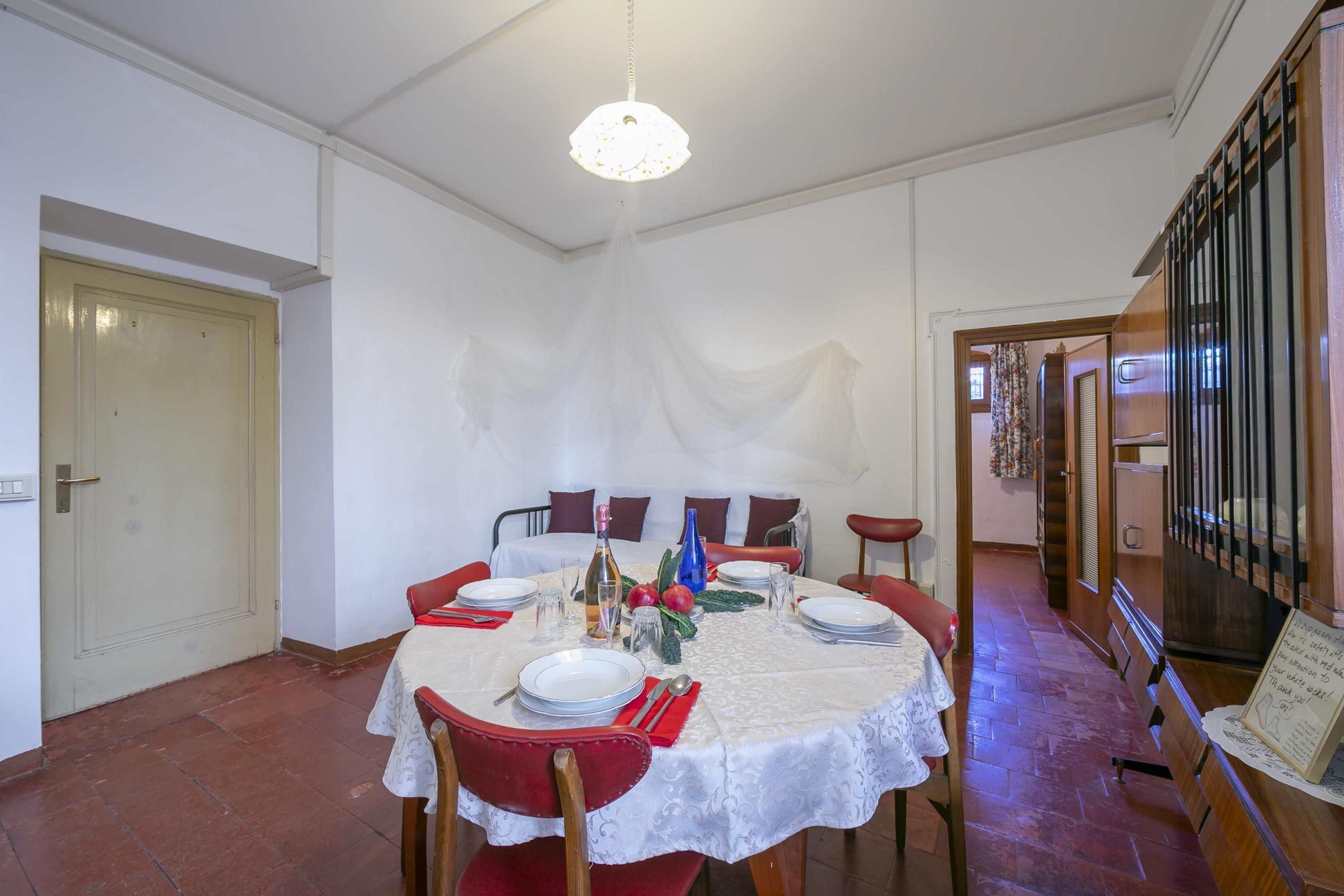 Een eetruimte met een ronde tafel voor vier personen, omringd door rode stoelen. Een kroonluchter hangt aan het plafond en een trap is rechts zichtbaar.