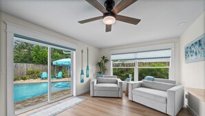 A spacious living room with a pool view, sliding glass doors, a ceiling fan, and a large window. - Island Oasis with Heated Pool, Putt Putt Course, Tiki Bar, and Beach Gear! (Merritt Island)