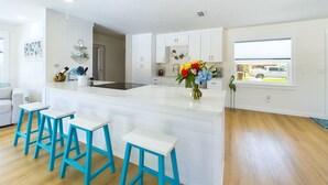 A modern kitchen with white cabinetry, a central island with stools, and a window overlooking a driveway. - Island Oasis with Heated Pool, Putt Putt Course, Tiki Bar, and Beach Gear! (Merritt Island)