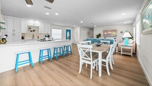A modern kitchen with white cabinets, a central island with blue stools, and a dining area with white chairs. - Island Oasis with Heated Pool, Putt Putt Course, Tiki Bar, and Beach Gear! (Merritt Island)