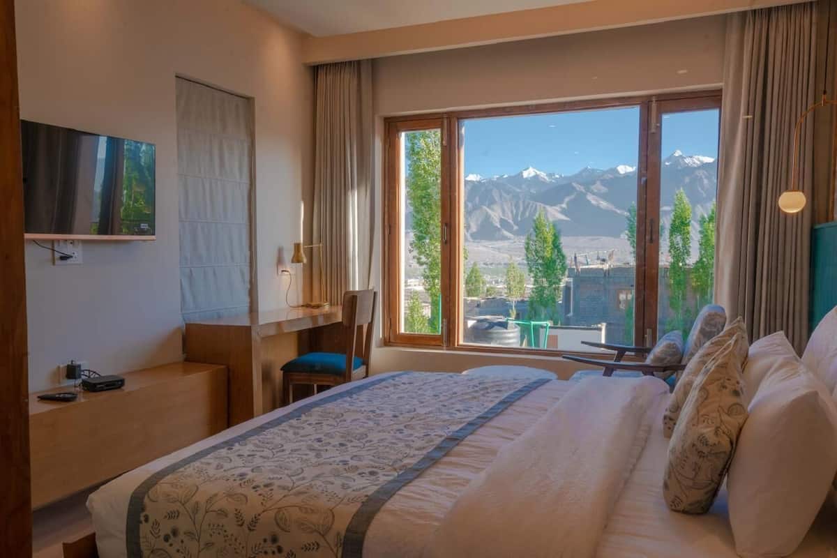 A hotel room with a large bed, a desk, a chair, and a TV. The room has a view of mountains through large windows.