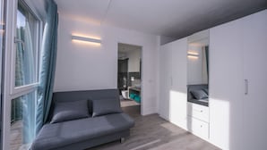 A modern bedroom with a grey sofa, white wardrobe, and a view through a window with curtains. - Rustichel Lake View in Toscolano Maderno (Toscolano Maderno)