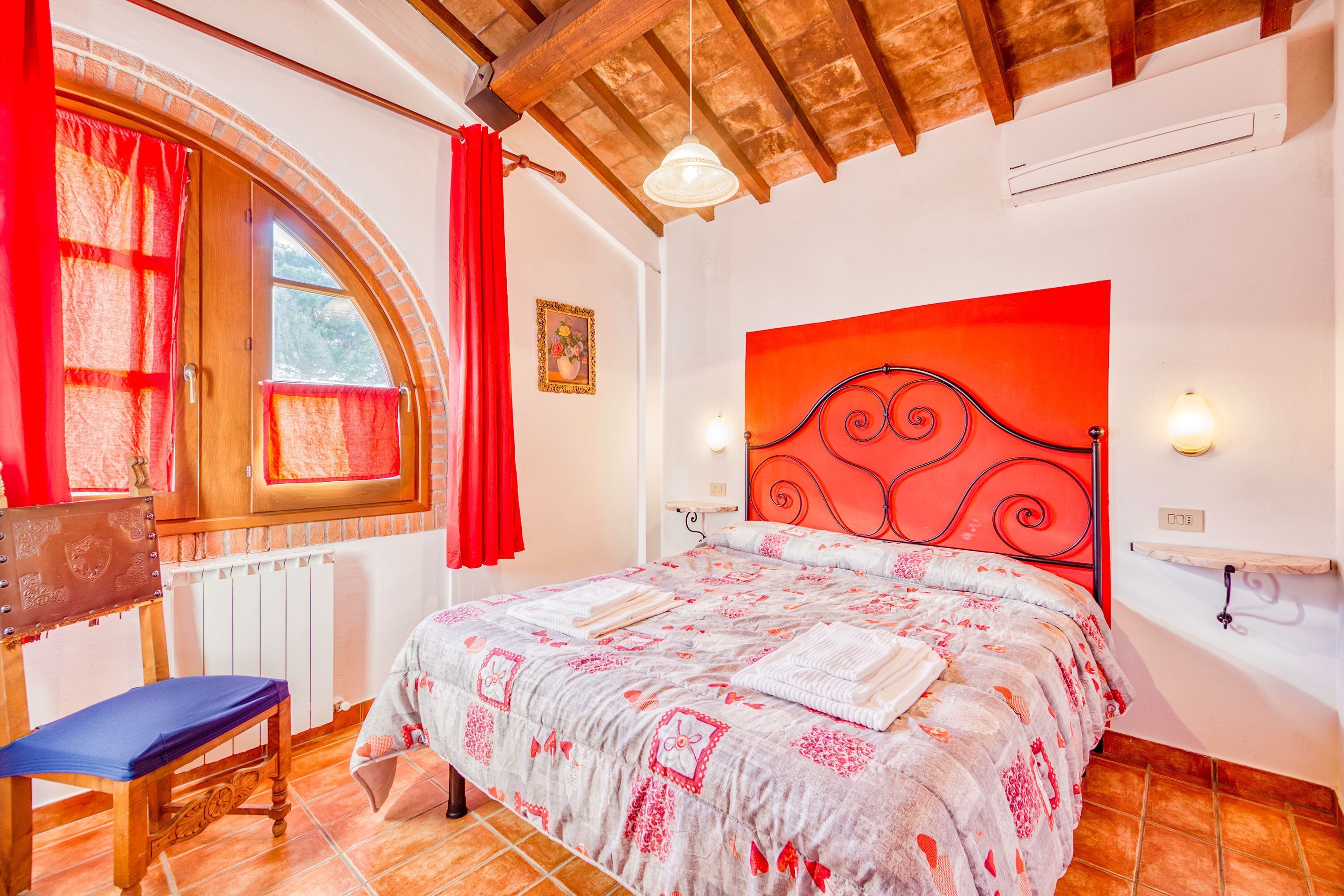 A bedroom with a large bed, a wooden bench, a window with red curtains, and a red headboard.